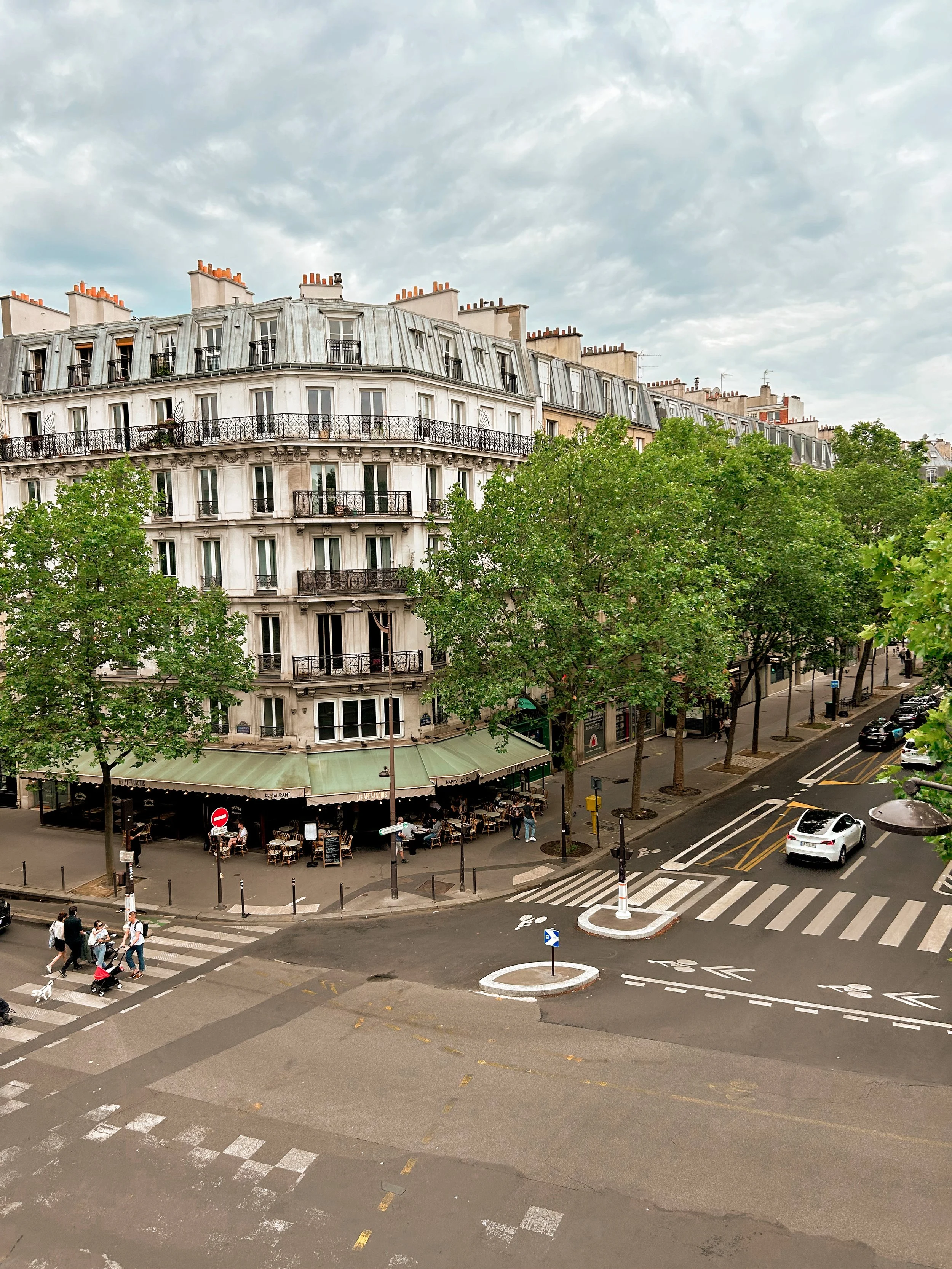 A street view in Paris showing an apartment building with curved balconies, lined with green trees, and a sidewalk café with outdoor seating. Cars are parked along the street, and pedestrians are crossing the crosswalk.