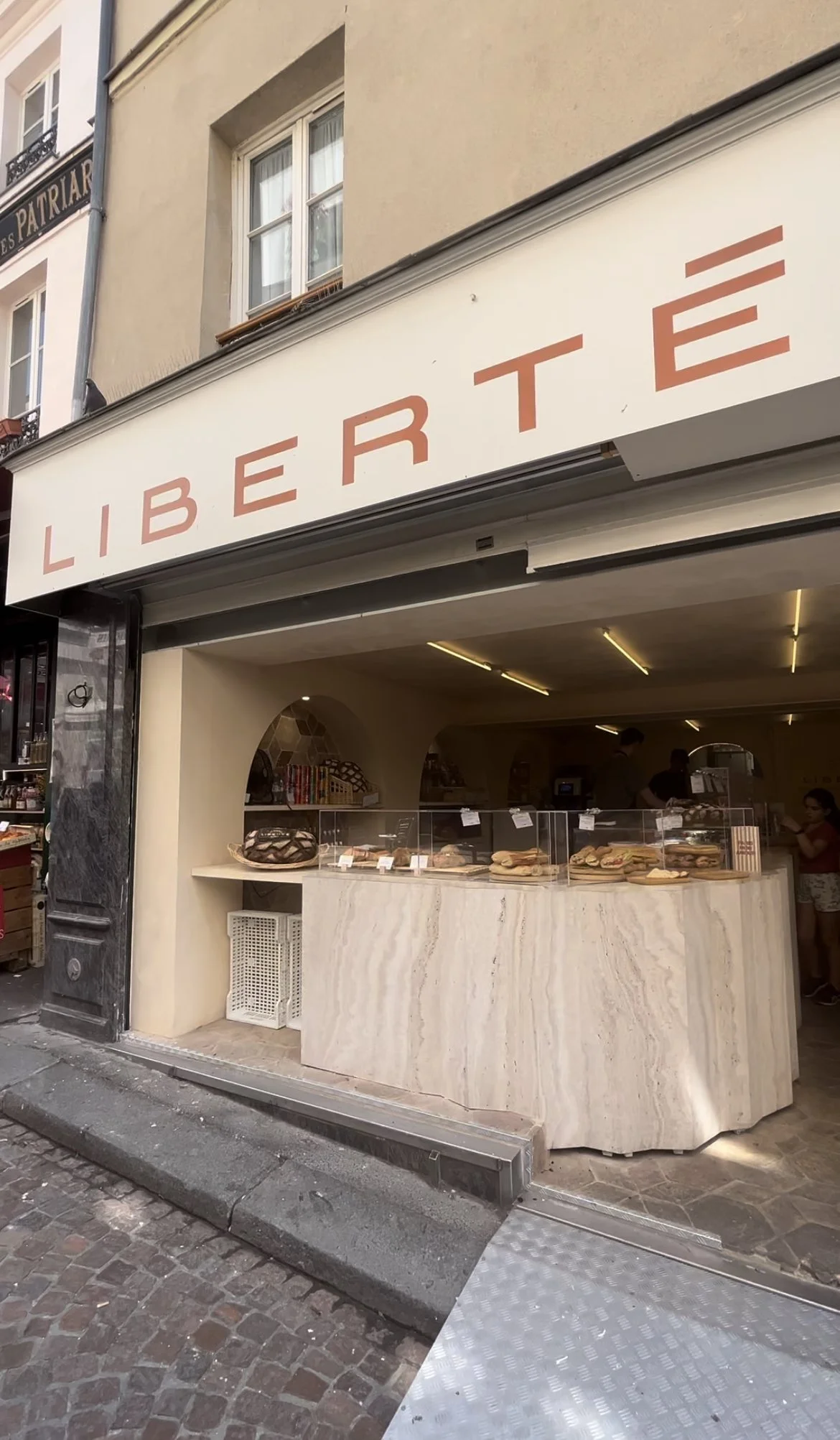 Exterior of a bakery or pastry shop with a sign that says 'LIBERTE' at the entrance, showing inside counter with pastries and baked goods, and a few customers inside.
