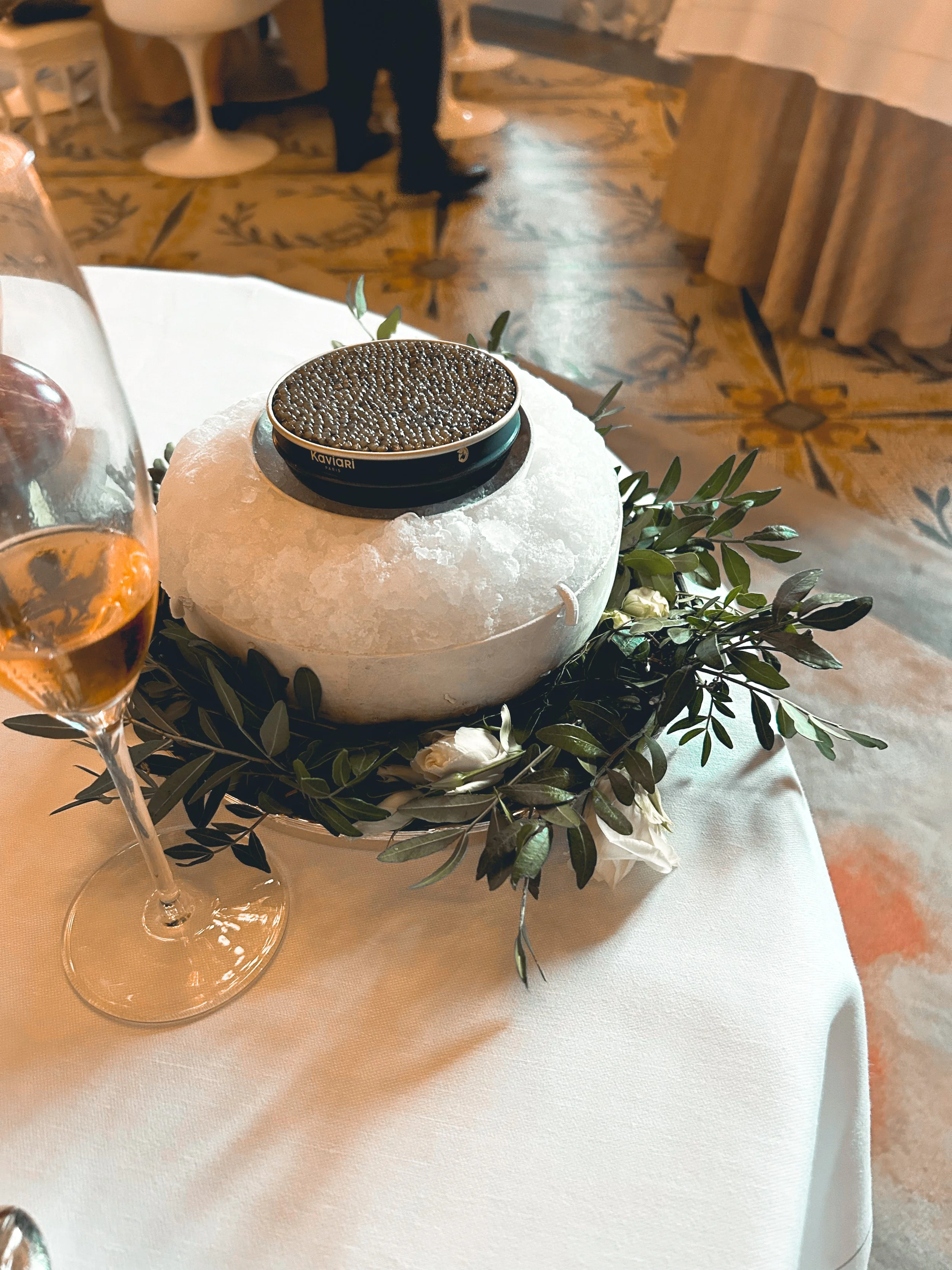 A round cheese wheel decorated with greenery and white flowers, with a small container of caviar on top and a glass of rosé wine nearby, on a white tablecloth.