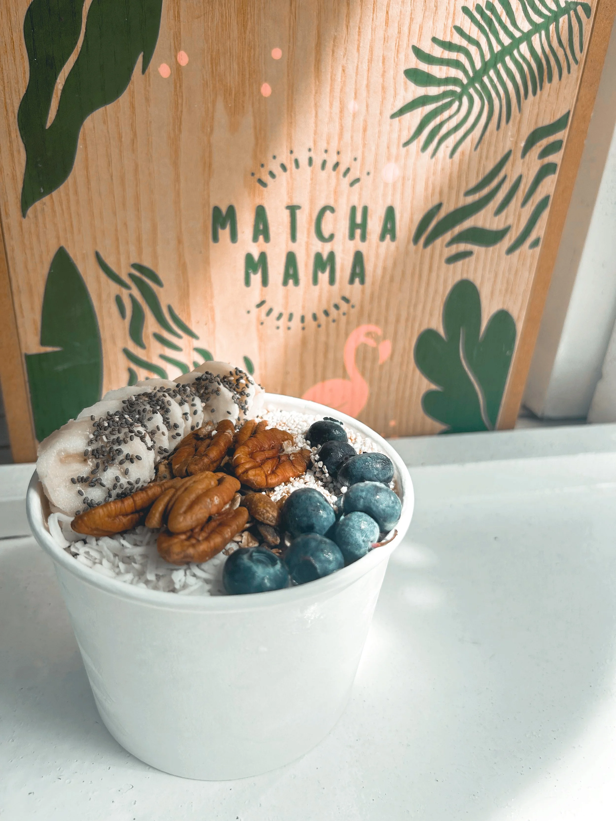 Potted matcha latte topped with blueberries, pecans, coconut flakes, and cookies with chocolate sprinkles, placed in front of a wooden sign that reads 'Matcha Mama' with botanical illustrations and a flamingo.