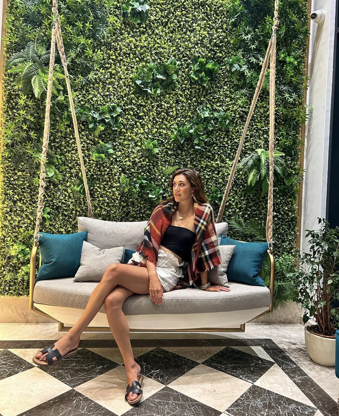 Woman sitting on a beige swing with colorful cushions, against a lush green plant wall background in an indoor setting.