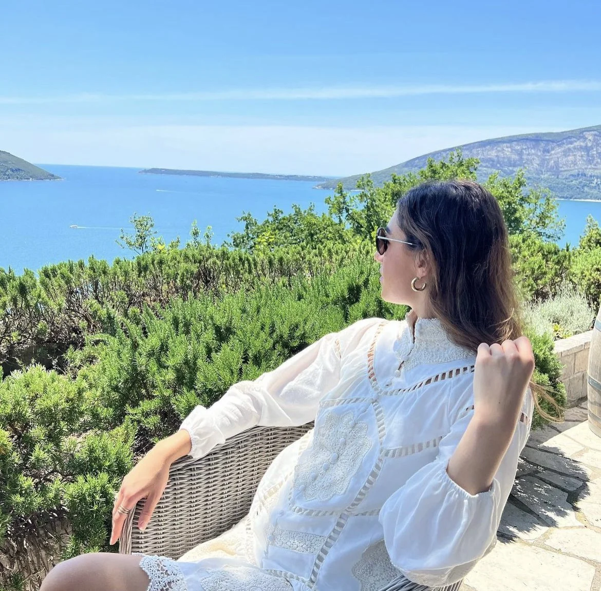 A woman with dark hair, wearing sunglasses, earrings, and a white embroidered blouse, relaxes outdoors on a wicker chair overlooking a scenic landscape of green shrubs, a blue lake, and distant hills under a clear blue sky.