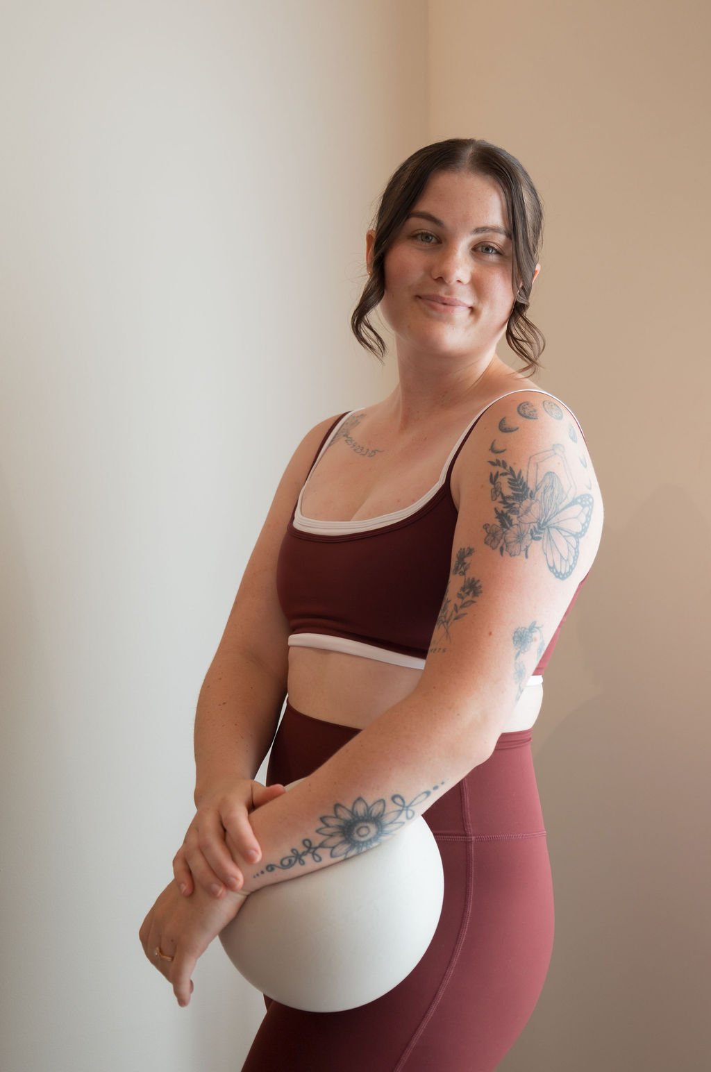 A young woman in activewear, holding a volleyball, standing indoors against a plain wall, smiling at the camera.