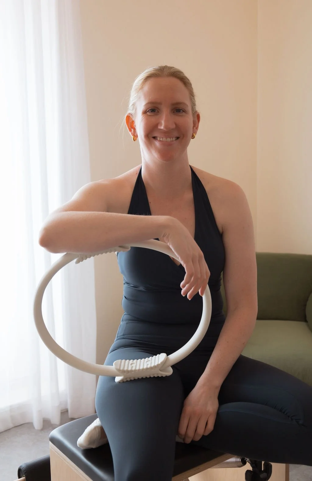 A woman with blonde hair smiling, wearing a black top, sitting on a medical examination table with a stethoscope draped over her arm, in a room with beige walls and a window with sheer curtains.