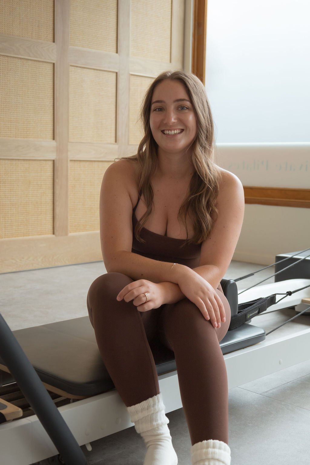 A woman with long light brown hair smiling at the camera, sitting on a Pilates reformer machine in a fitness studio with beige and wooden decor.