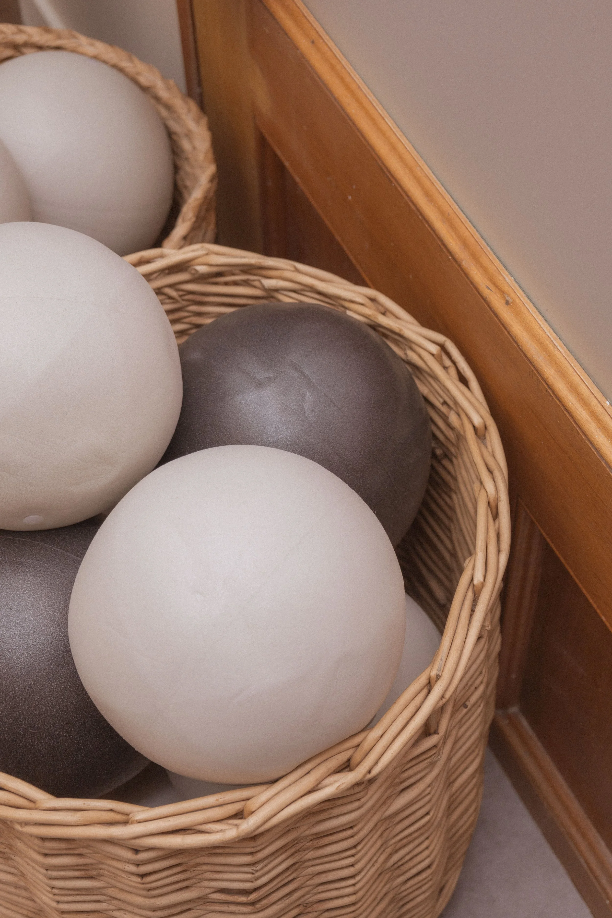 A woven basket containing large white and gray balls, placed next to a wooden piece of furniture and a wall.