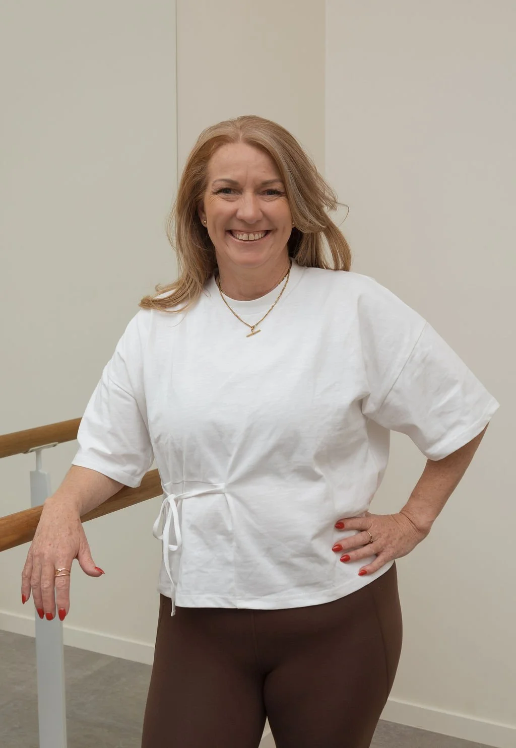 Woman in white top and brown pants smiling, standing next to a wooden railing in an indoor setting.