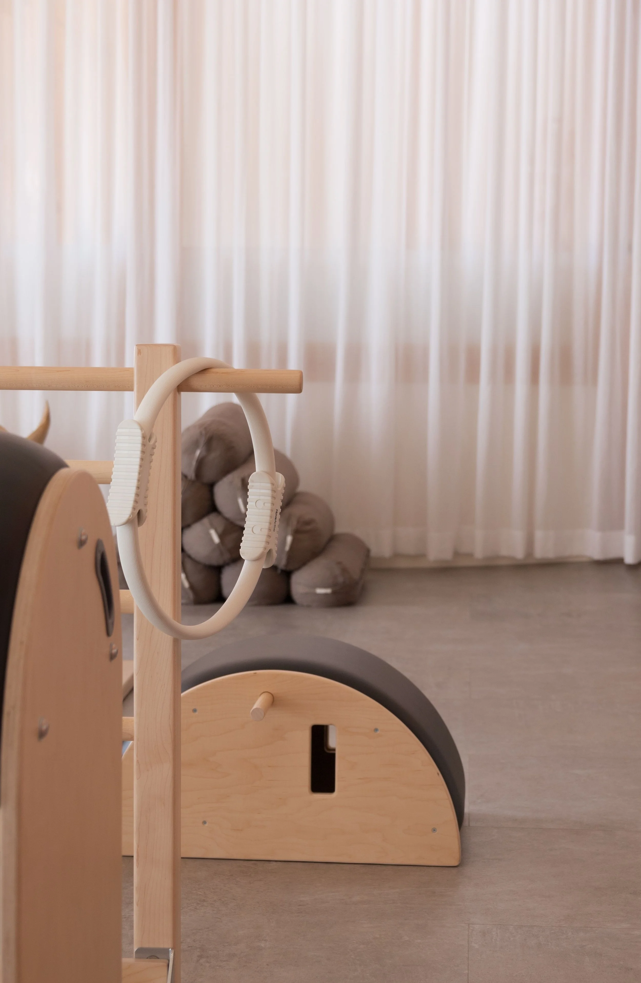 A wooden therapy or exercise equipment piece with a curved black roller and a white hand grip attachment, set on a light-colored floor with a backdrop of stacked brown fabric cushions and white curtains.