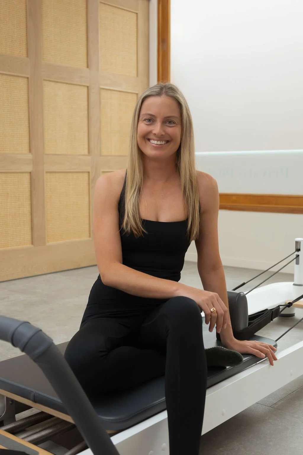 A woman sitting on a Pilates reformer machine in a fitness studio, smiling at the camera, wearing a black tank top and black pants.