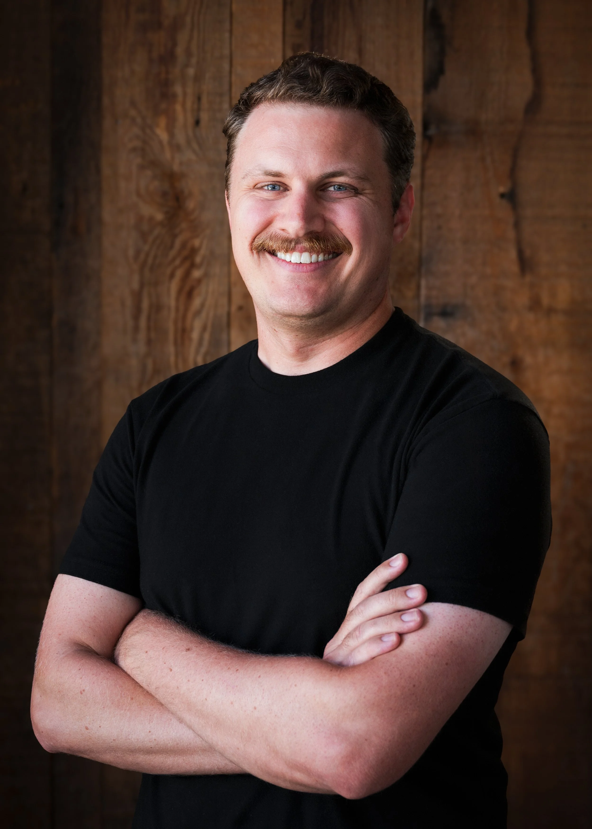 A smiling man with a mustache, crossed arms, wearing a black t-shirt, standing in front of a wooden background.