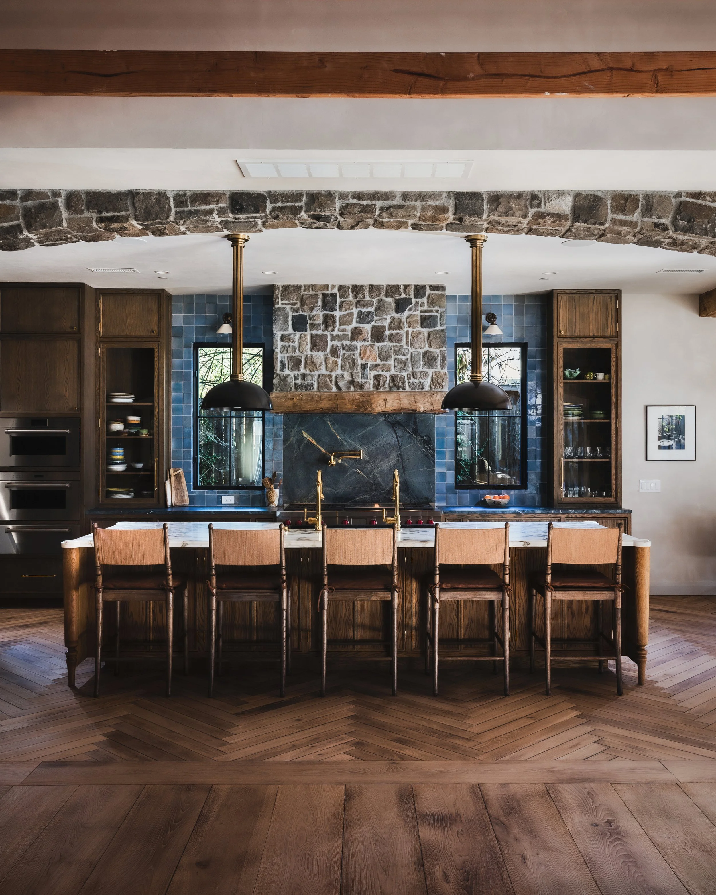 Interior of a modern kitchen with a large wooden dining table, eight wooden chairs, a brick and stone fireplace, dark wooden cabinets, blue tiled backsplash, and black pendant lights hanging from the ceiling.