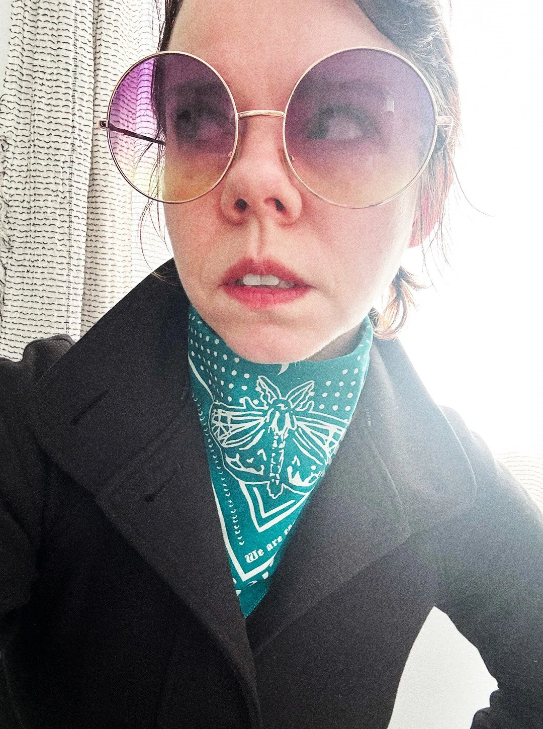 Woman looks irritated and looks to the side wearing oversized round lens sunglasses, black goth jacket, and a teal bandana that showcases a large moth on the point.