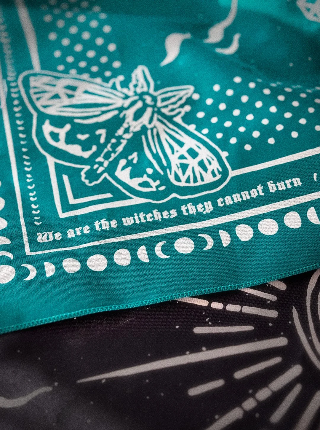 close up of Aqua Seas colored Sugar Camp bandana on distressed wood floor with moth corner detail, polka dots around the edge, and the saying "we are the witches they cannot burn"