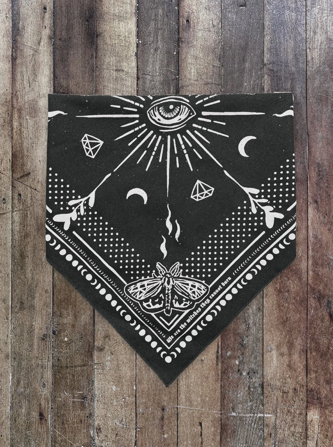 Folded raven black Sugar Camp bandana on distressed wood floor with moth corner detail, an eye in the center, polka dots around the edge, and the saying "we are the witches they cannot burn"
