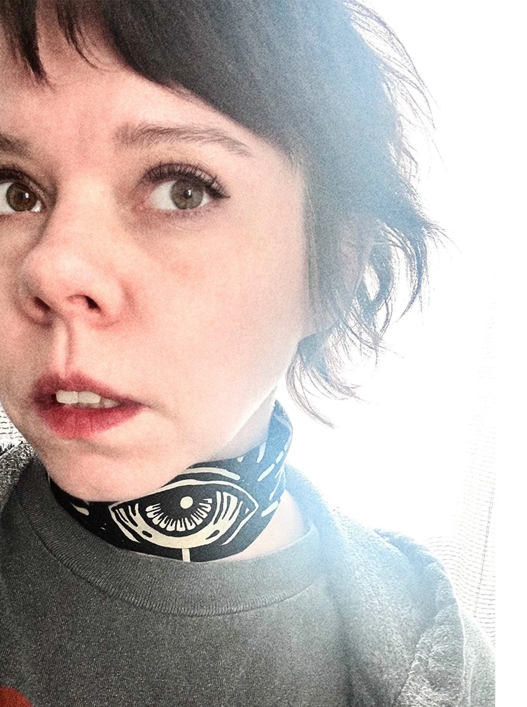 Side of the face of a woman with short brown hair and red lipstick wears a black bandana rolled around neck that shows a large "third-eye" graphic in center.