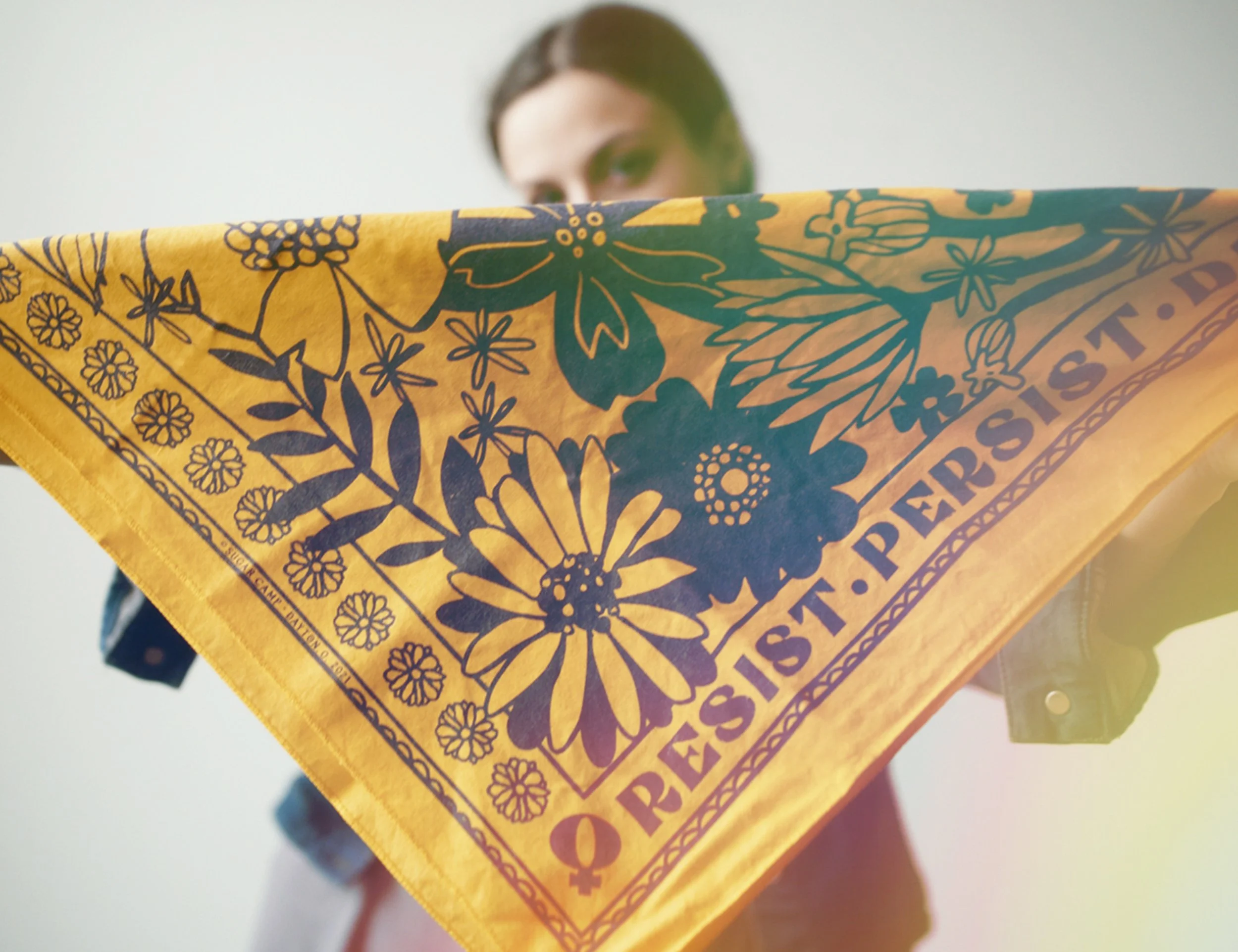 woman holds gold & navy resist floral bandana from the corners. by sugar camp.