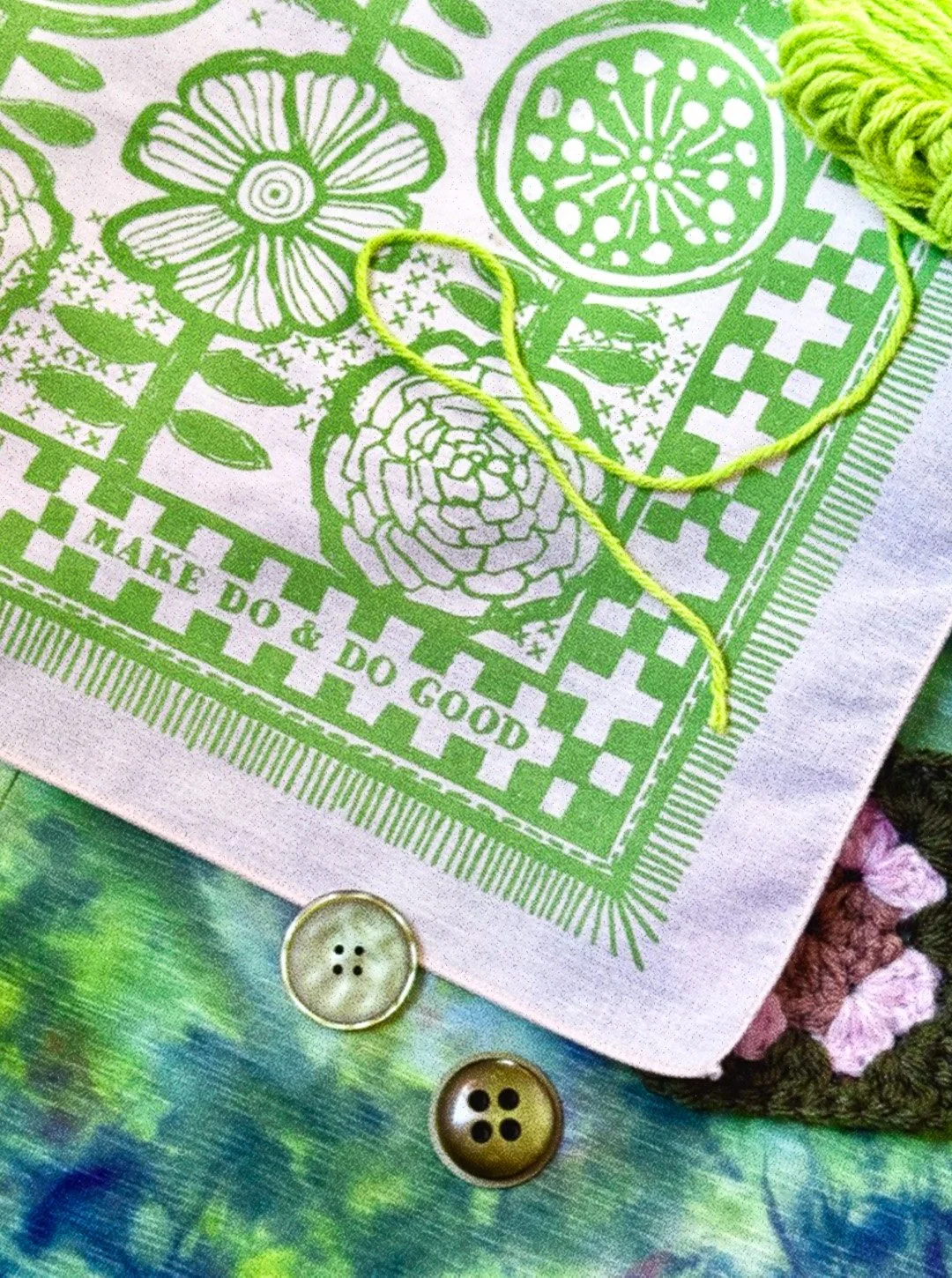 Sugar Camp cream bandanna with sprout green ink features hand-drawn Scandinavian florals & stitches, reads “Make do & Do Good.” staged with with yarn and buttons