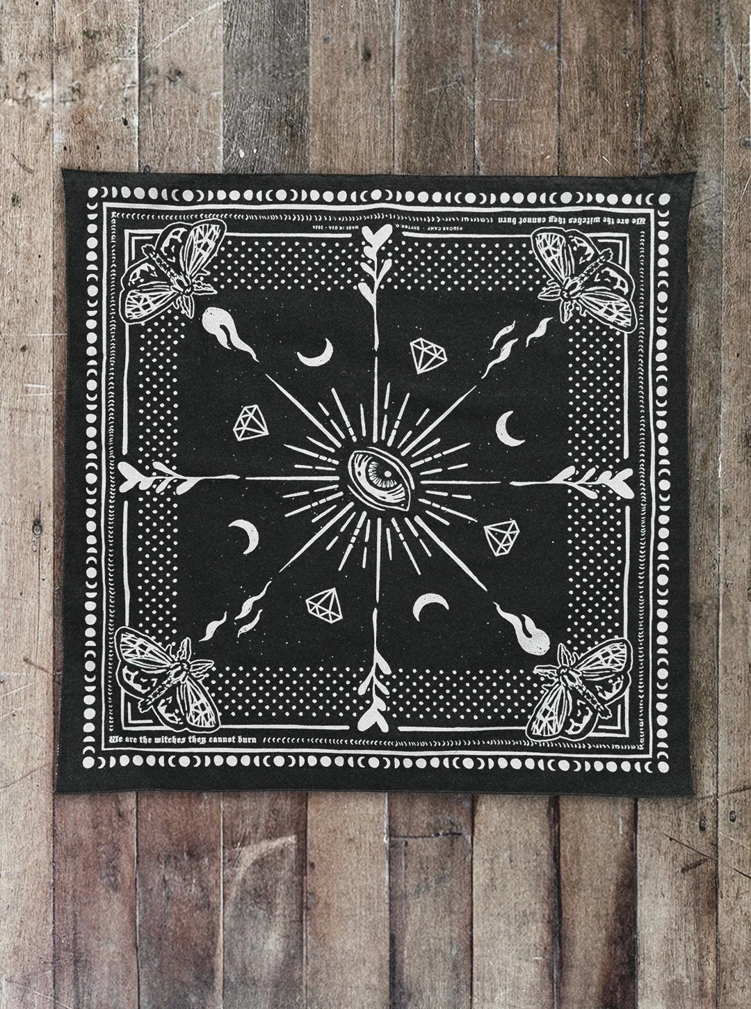 raven black Sugar Camp bandana laid flat on distressed wood floor with moth corner detail, an eye in the center, polka dots around the edge, and the saying "we are the witches they cannot burn"