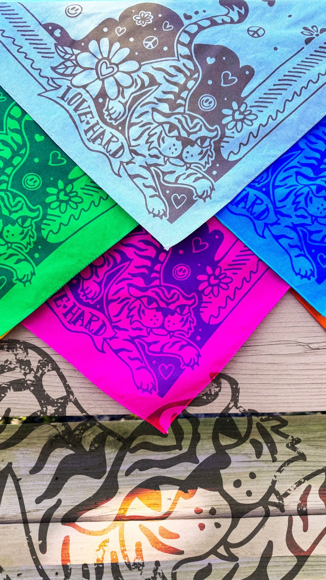Love Hard Bandana rainbow collection by Sugar Camp in a spectrum of colors and ink, stacked on a weathered wood background. Features sweet, fierce tiger, hearts, smileys, and peace signs.