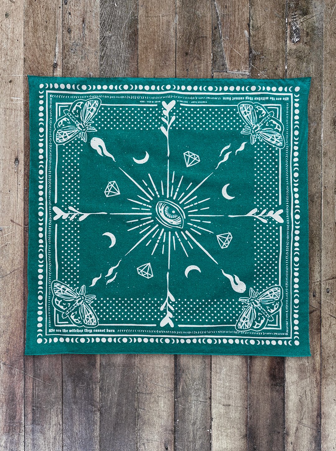 Aqua Seas-colored Sugar Camp bandana laid flat on distressed wood floor with moth corner detail, an eye in the center, polka dots around the edge, and the saying "we are the witches they cannot burn"