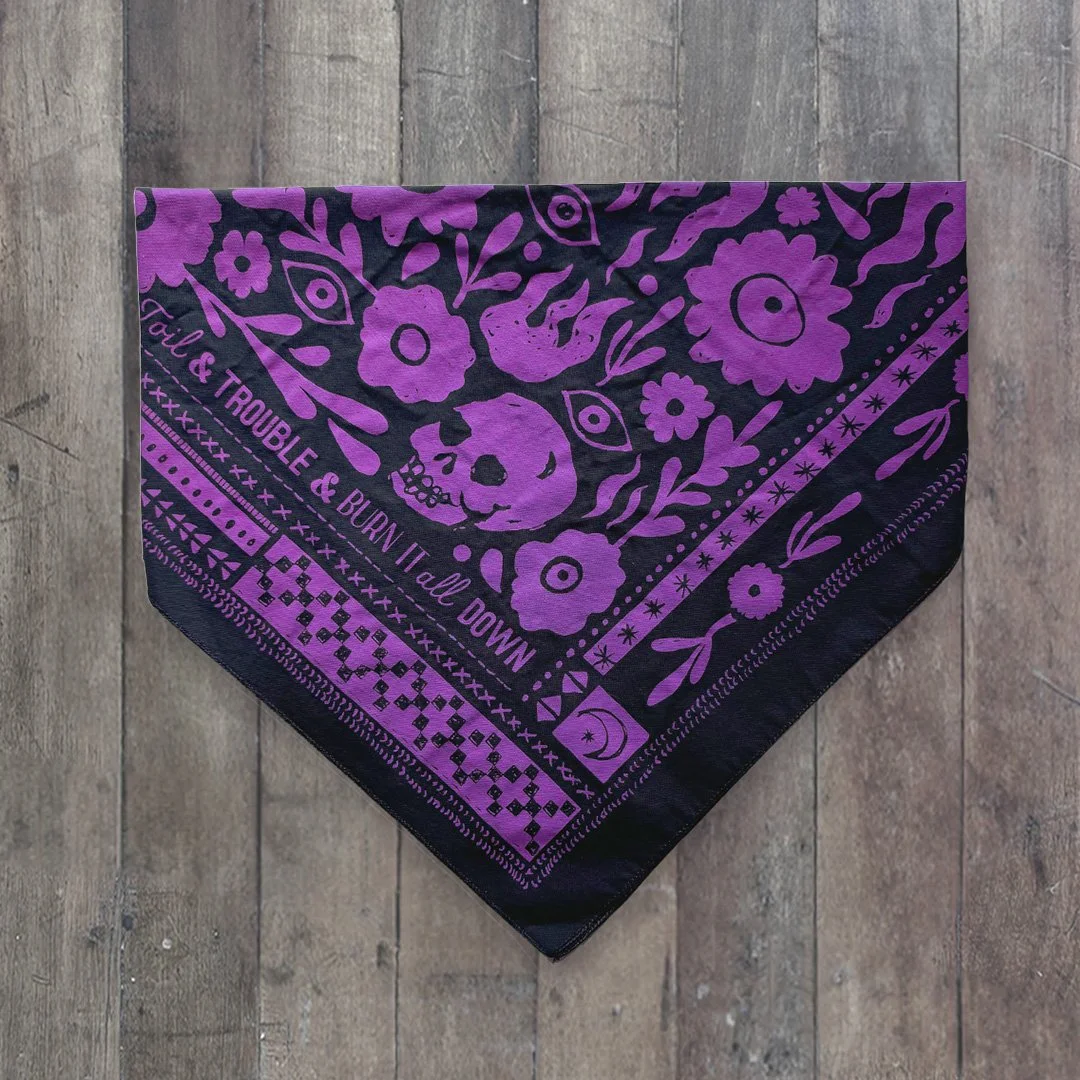 Toil & Trouble Bandana by Sugar Camp, in Violent Violet color palette. Folded on worn wood floor.
