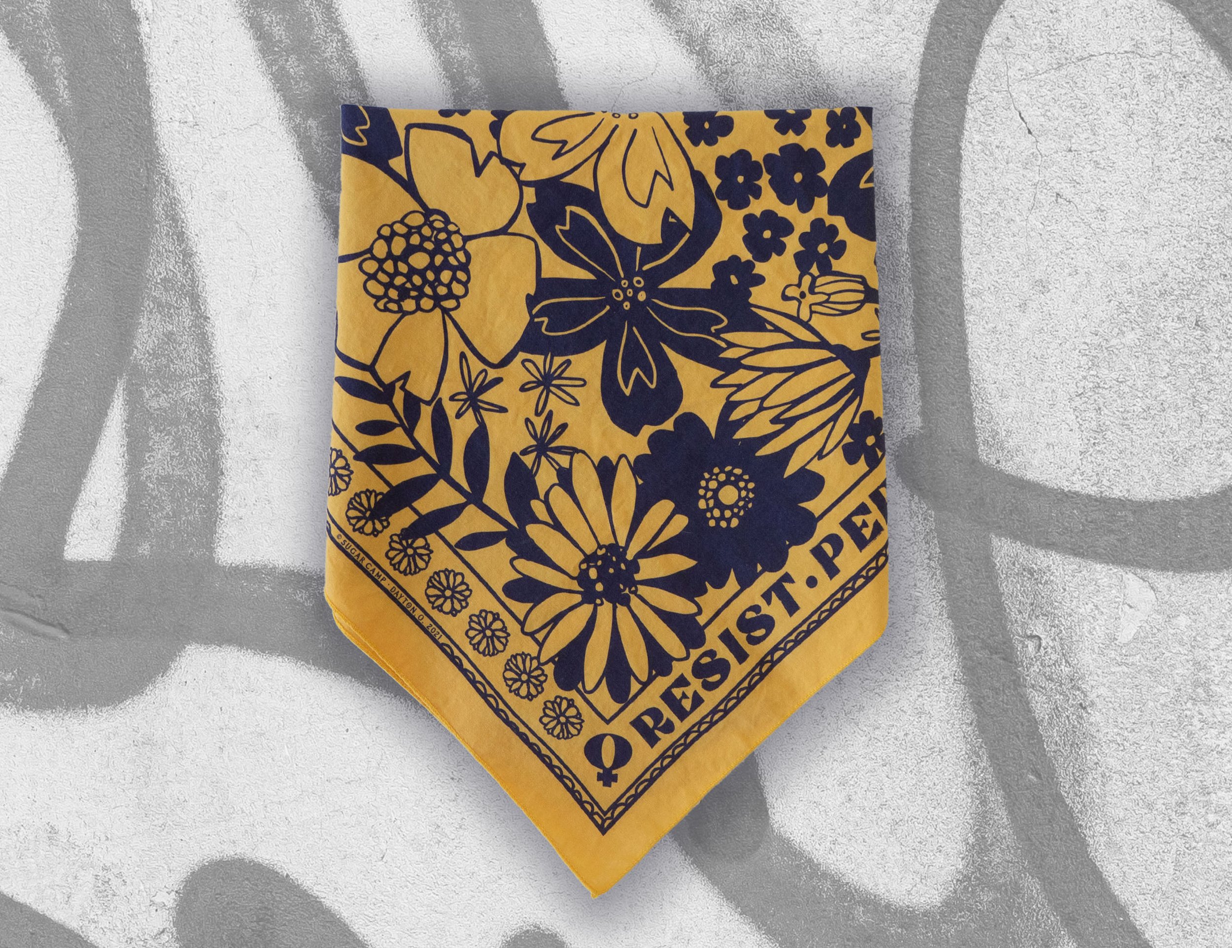 Folded mustard yellow floral bandana with bold 'RESIST' text, inspired by Elizabeth Warren, Ruth Bader Ginsburg, and Rosa Parks — feminist empowerment accessory promoting social justice and equality.
