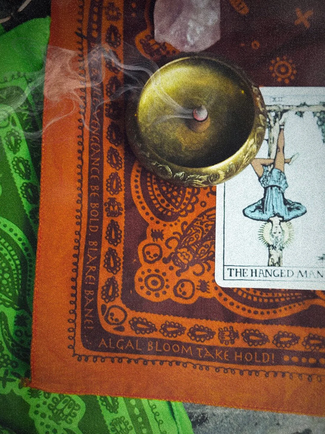 burnt orange swamp curse bandana flat lay with paisley print and algal hex. The hanged man tarot and an incense cone sit on top of bandana.
