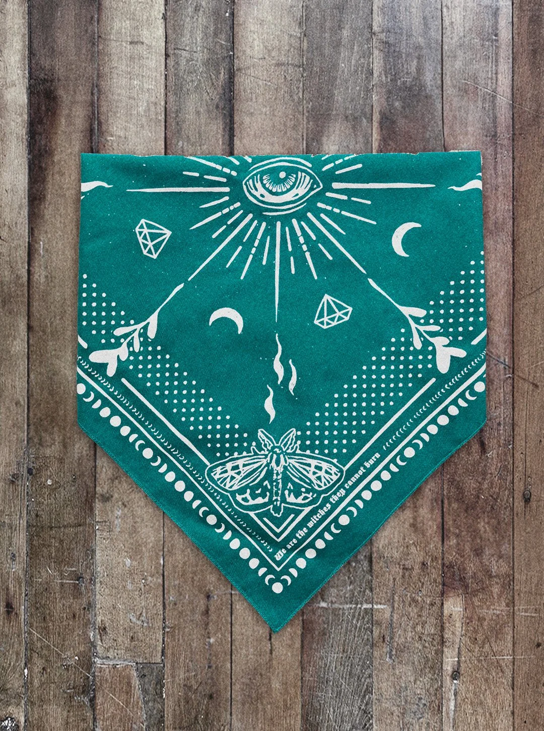 Folded Aqua Seas colored Sugar Camp bandana on distressed wood floor with moth corner detail, an eye in the center, polka dots around the edge, and the saying "we are the witches they cannot burn"