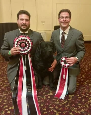 Two men in suits kneeling with a black retriever, holding award ribbons, in a decorated room