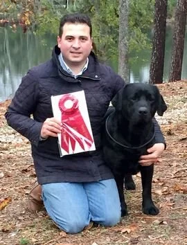 A man kneeling outdoors next to a large black dog near a body of water with trees in the background. The man is holding a piece of paper or a sign.