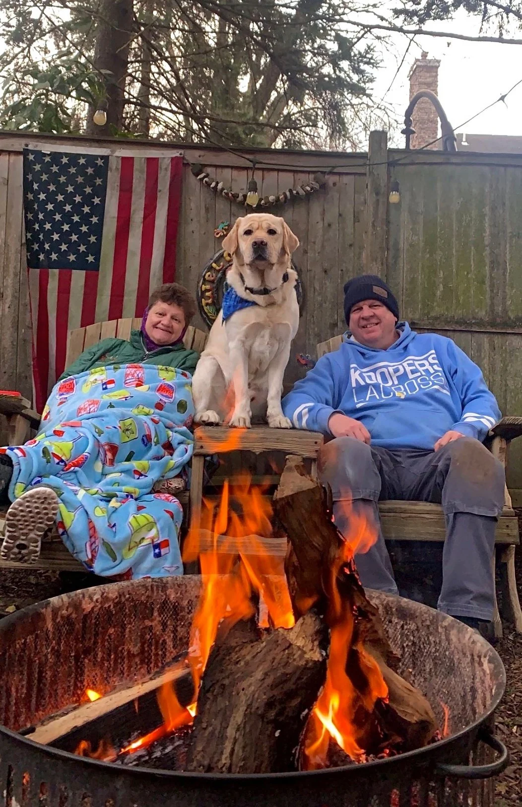 A woman and a man sitting on outdoor chairs by a fire pit, with a large yellow labrador retriever standing on the backrest between them. The woman is wrapped in a colorful blanket, and the man is wearing a blue hoodie. There is an American flag hanging on the fence in the background and a string of lights above.