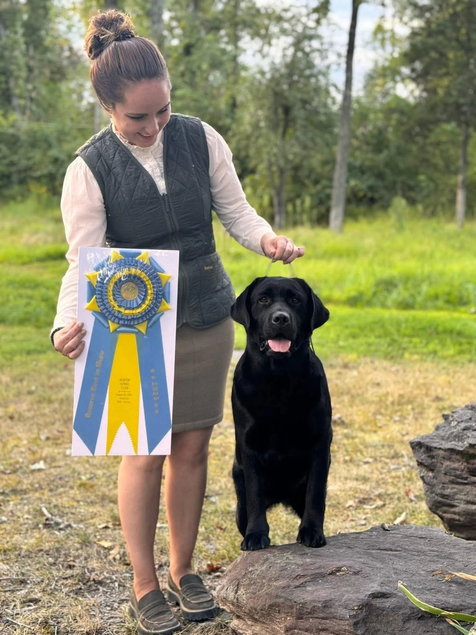 A woman with a bun hairstyle is standing next to a black Labrador Retriever on a rock, holding its leash. She is smiling and holding a blue and yellow rosette ribbon award in her left hand. They are outdoors in a wooded area, celebrating the dog's ac