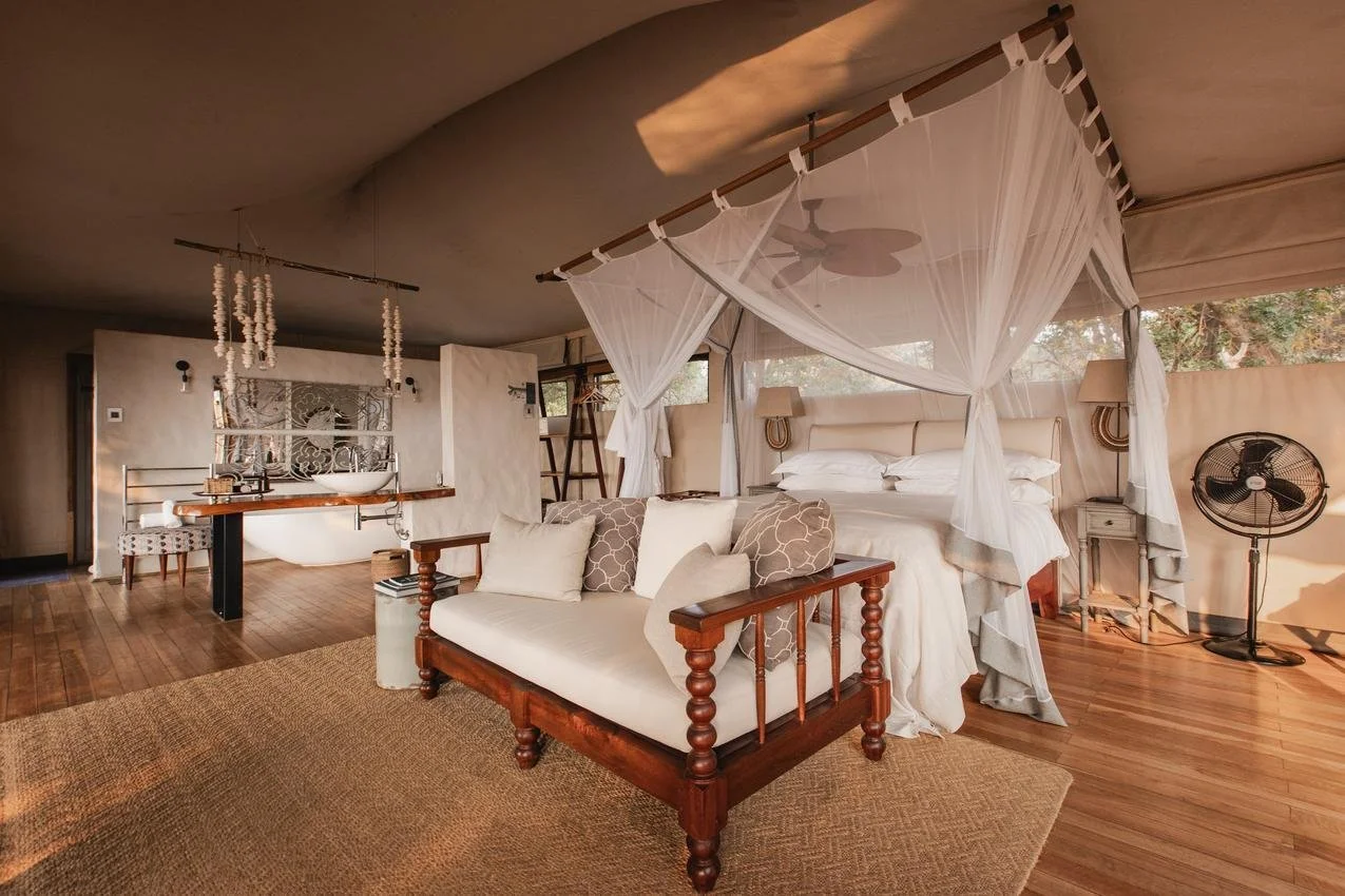 A cozy bedroom with a four-poster bed draped in white curtains, located in a warm-toned room with hardwood floors. There is a small white sofa with throw pillows in front of the bed, a standing fan on the right, and a small nightstand. In the backgro