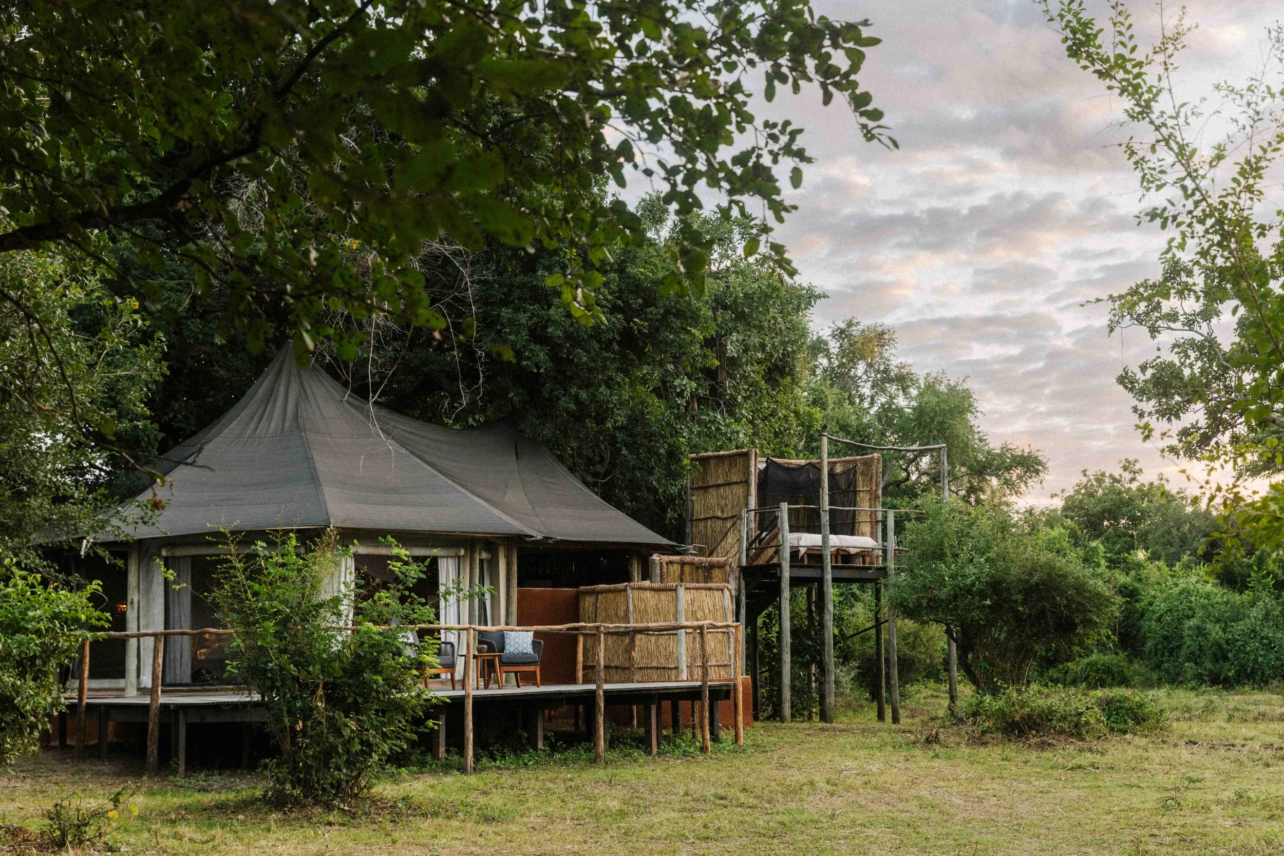 A rustic lodge or cabin with a thatched roof, sitting on stilts amidst greenery. There is a small porch with chairs, and next to it is an elevated platform or deck with a bed, surrounded by wooden railings, set in a natural outdoor environment.