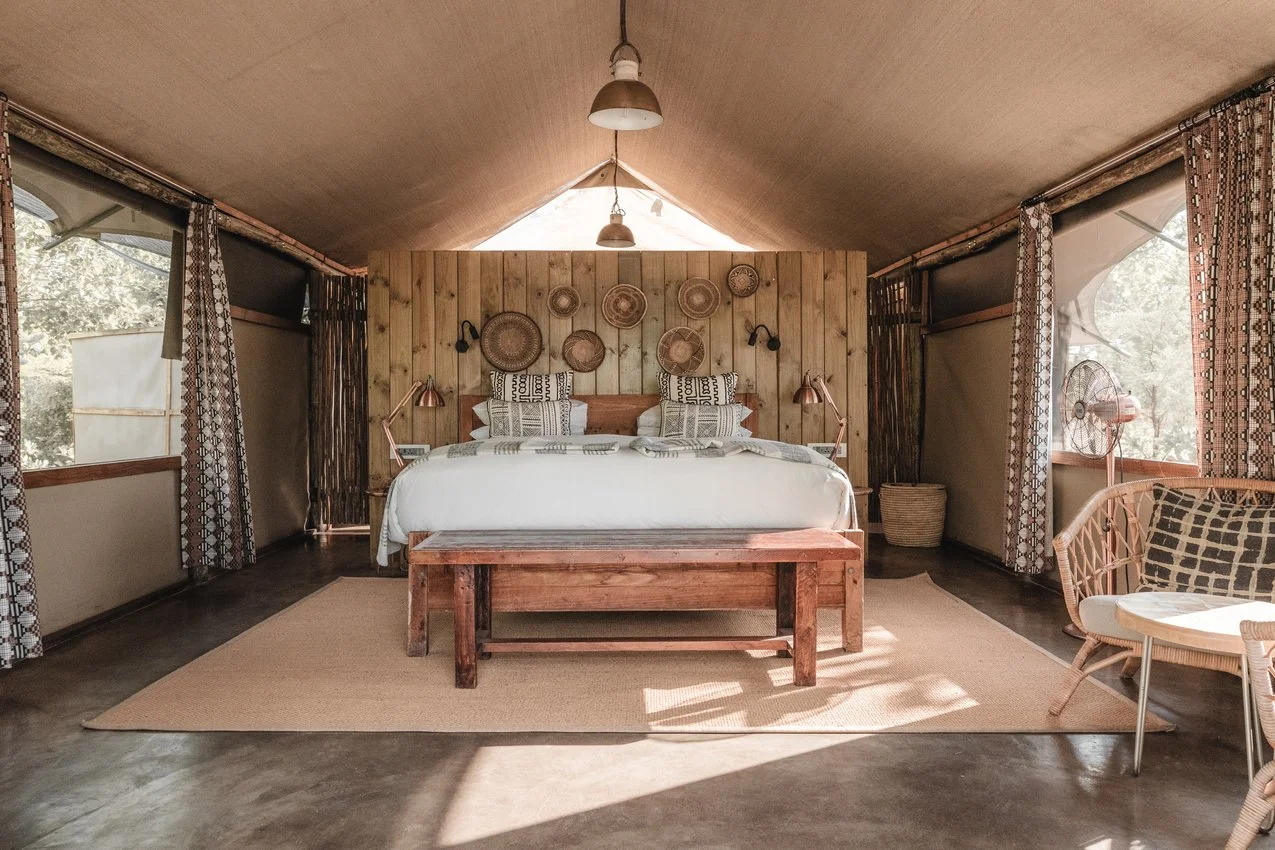 Clean, rustic bedroom with a wooden headboard, decorative woven baskets on the wall, white bedding, and side lamps. Windows have patterned curtains, and there is a wooden bench at the foot of the bed and a seating area with wicker furniture on the ri