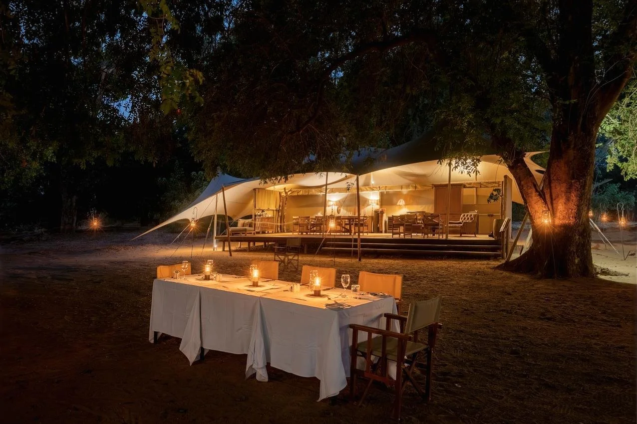 A large outdoor dining tent illuminated with warm lights, set up on a sandy ground with trees around, and a table with chairs and candlelit decorations in the foreground.