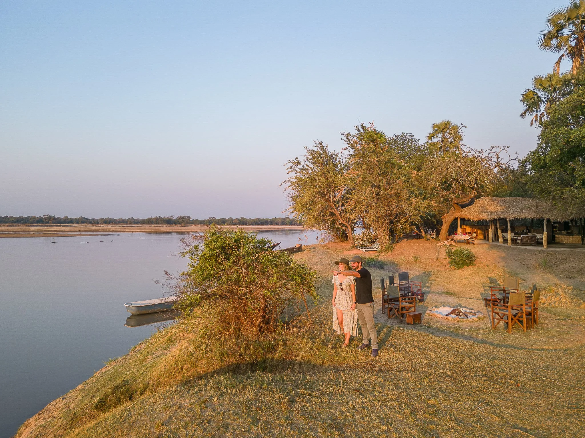 A couple standing by a riverbank at sunset, with chairs arranged around a fire pit and a thatched-roof structure in the background, surrounded by trees and greenery.