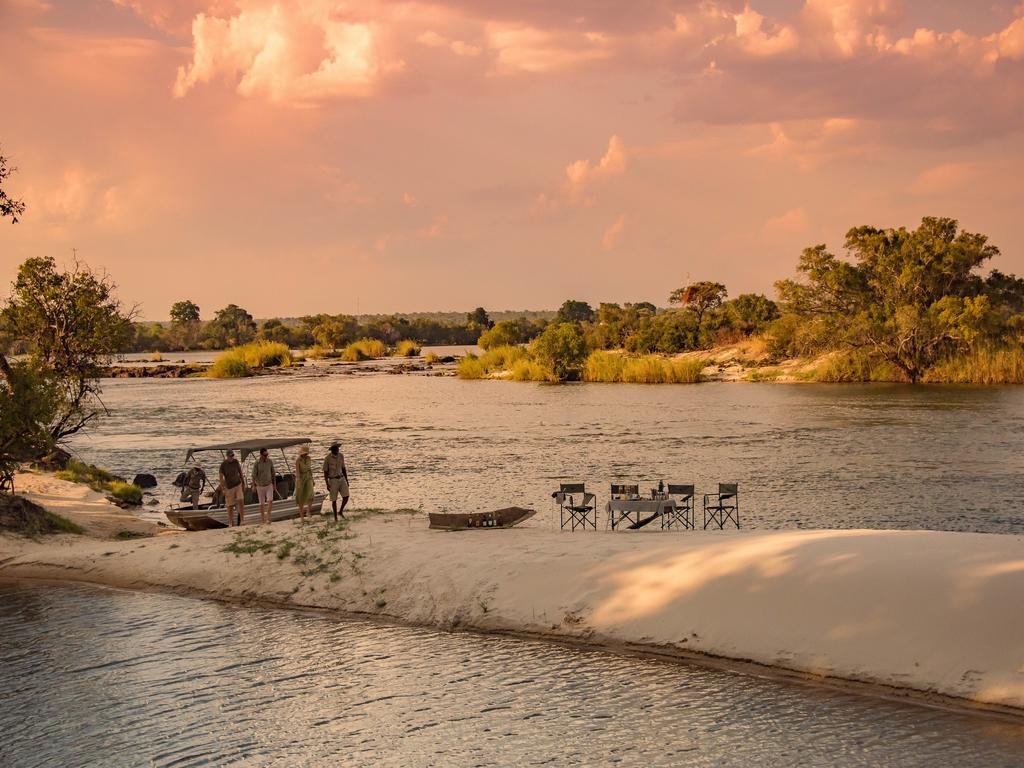 Group of people preparing to board a boat on riverbank during sunset, with outdoor dining tables set nearby and lush trees in the background.