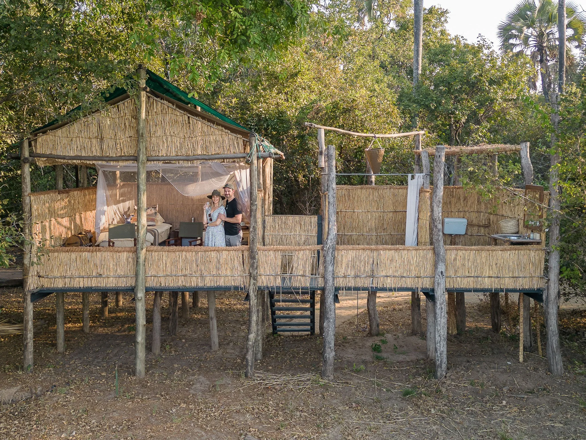 A rustic treehouse with a bamboo railing, containing a bed, small table, and chairs, with two people inside, and surrounded by lush greenery.