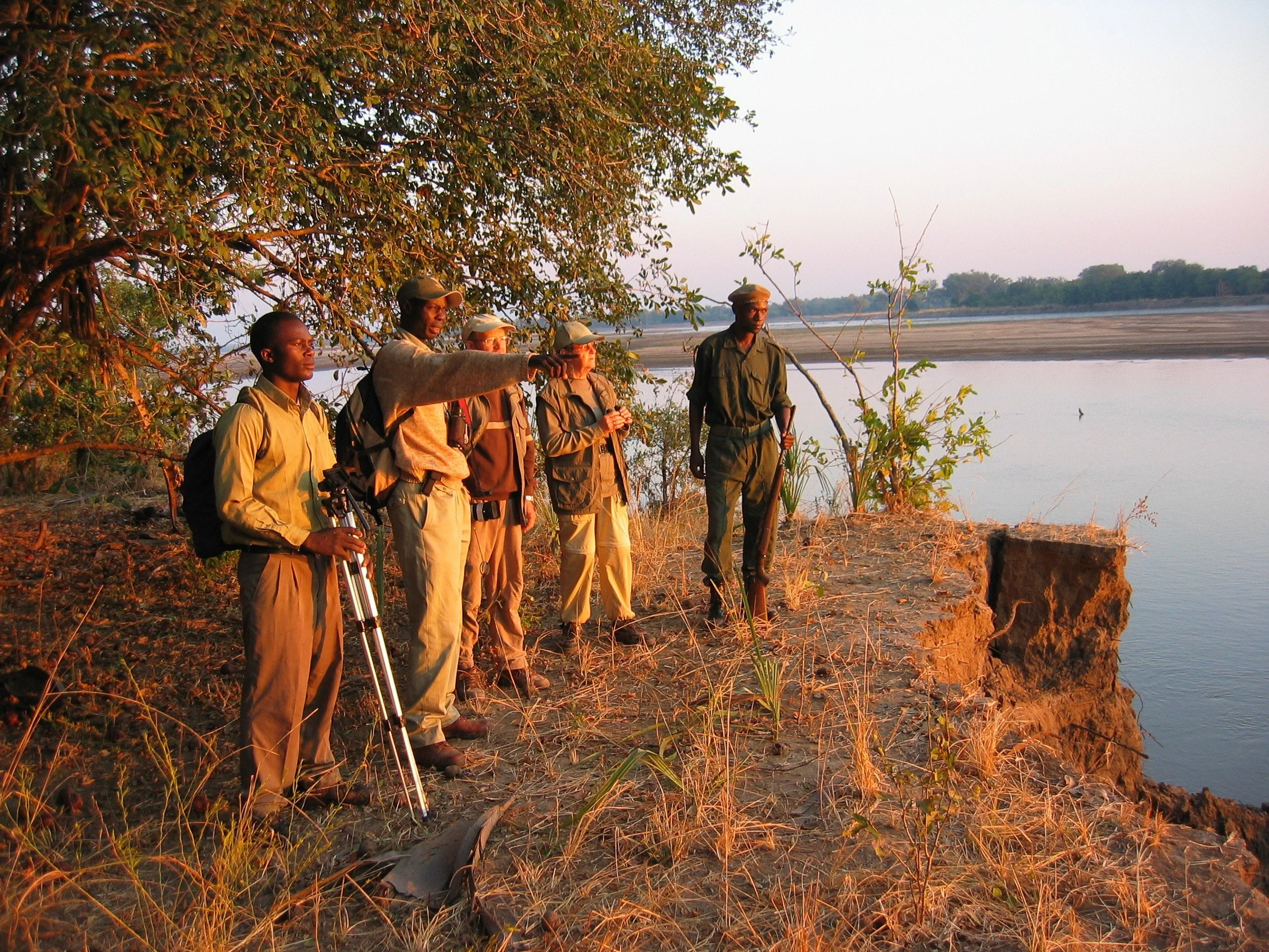 Group of six men outdoors near a body of water, one pointing into the distance, during sunset or sunrise.