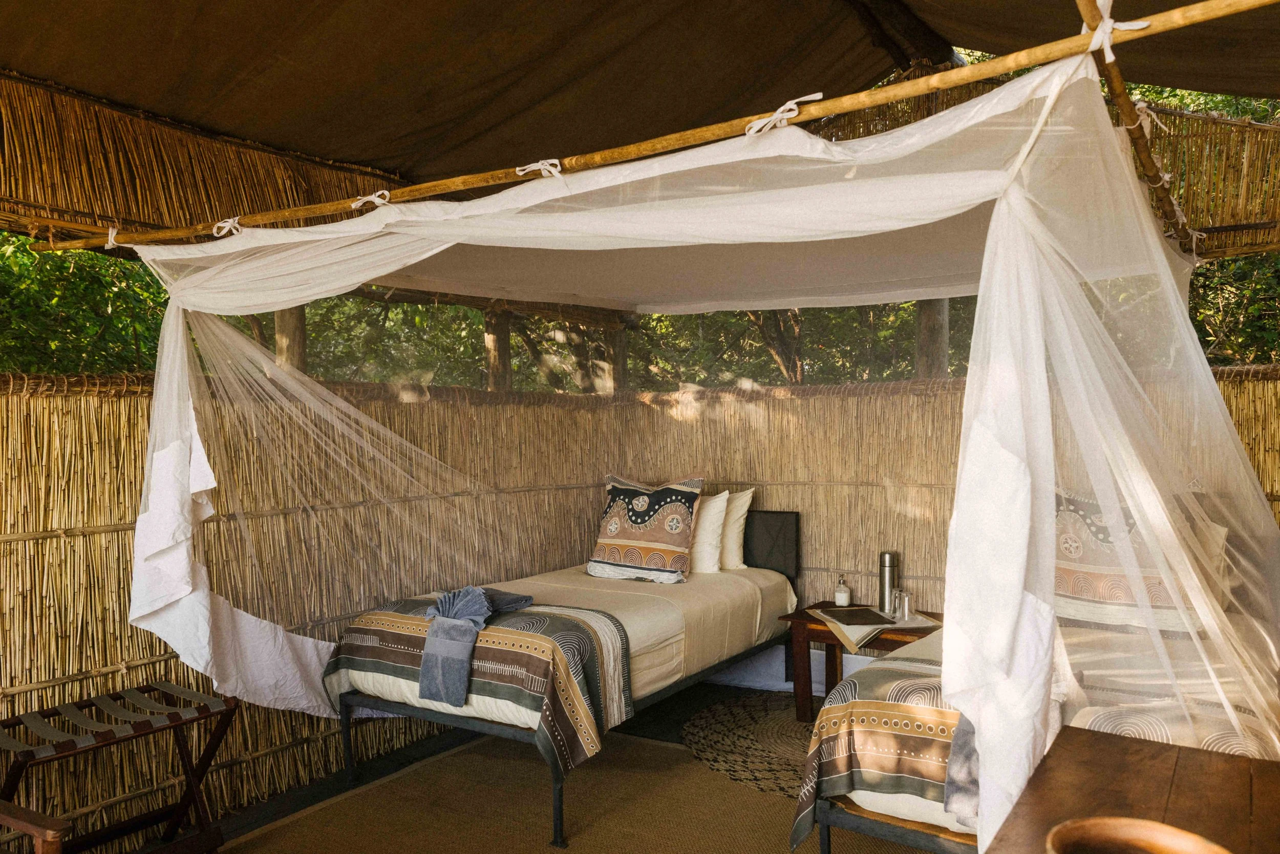 A cozy outdoor bamboo cabana with a canopy, featuring two beds with patterned bedding and pillows, surrounded by woods, and a small wooden side table with a book, thermos, and water bottle.