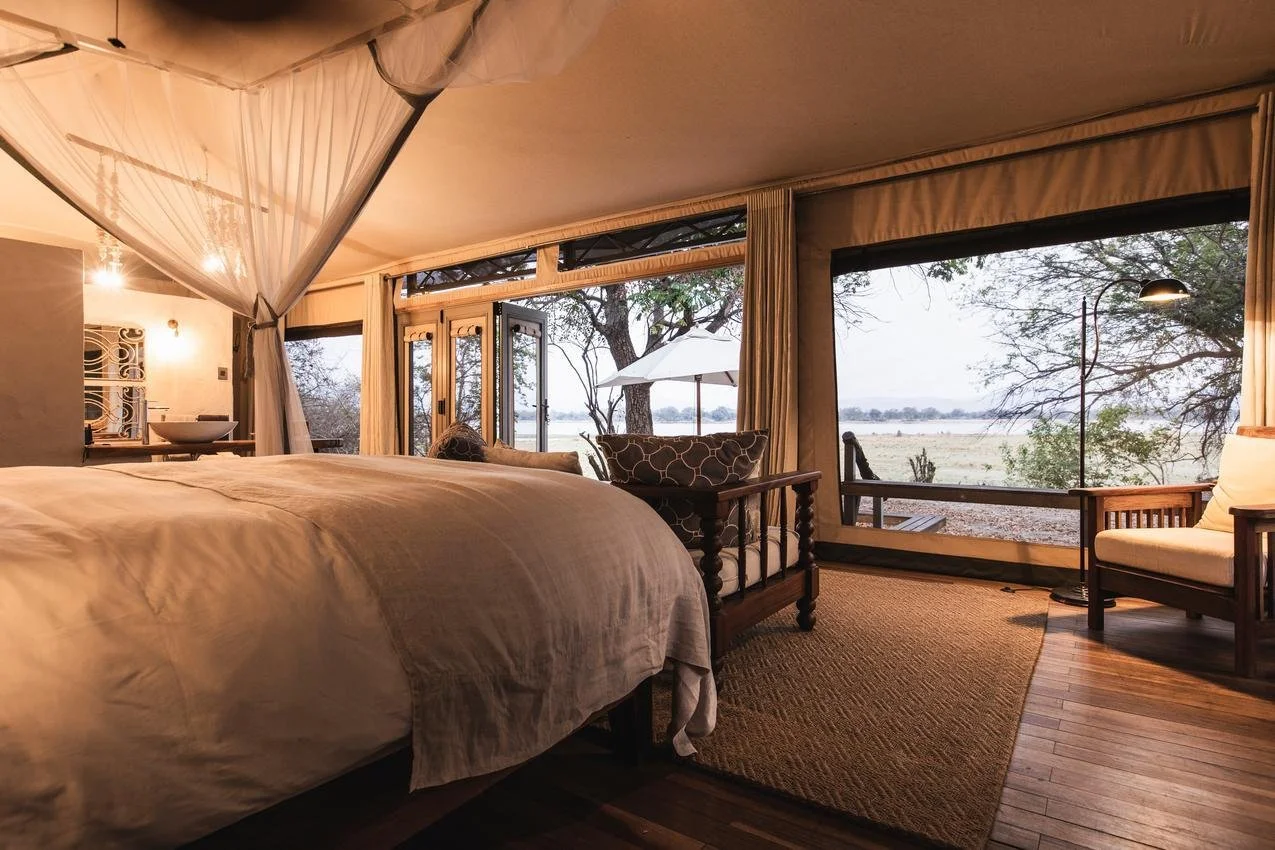A cozy bedroom with large windows overlooking an outdoor landscape with trees and a body of water, decorated with neutral tones and wooden furniture.