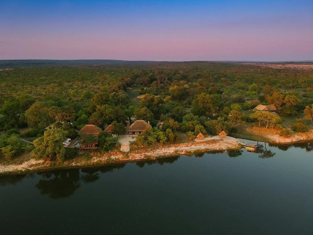 Aerial view of a lakeside area with trees, small huts, and a boat dock, at sunset with a hazy sky in the background.