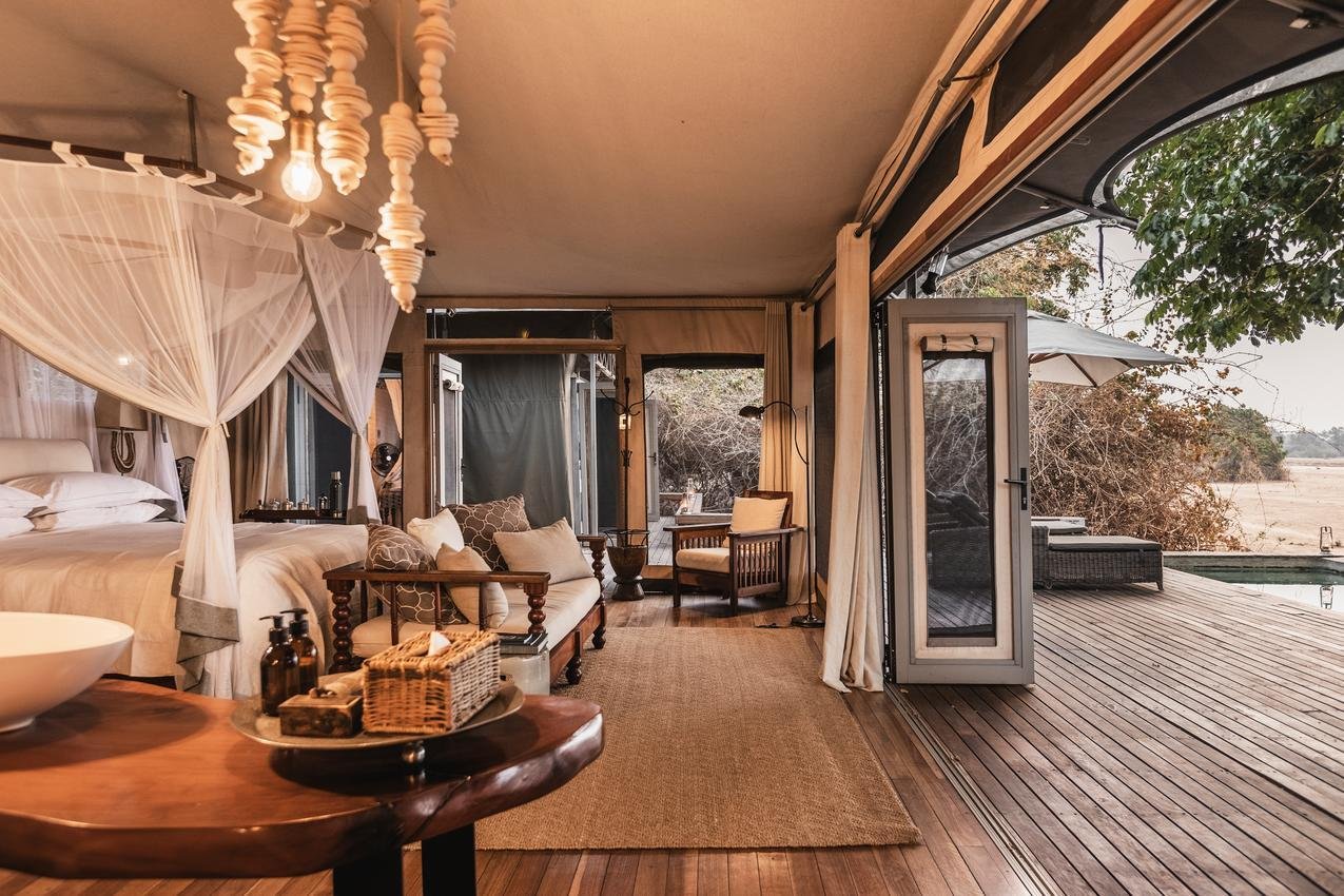Interior of a luxury tent with a bed, sofa, and seating area, opening to a wooden deck with outdoor furniture and a view of trees and fields.