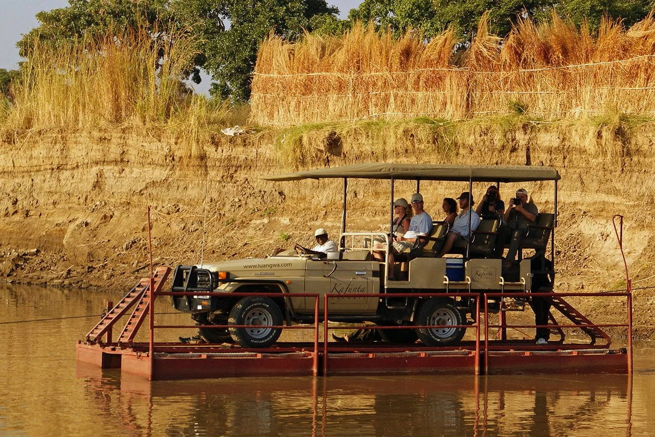 A tourist safari vehicle float on water with several passengers, some taking photos, passing along a riverbank with grassy and dirt embankment layered with trees.