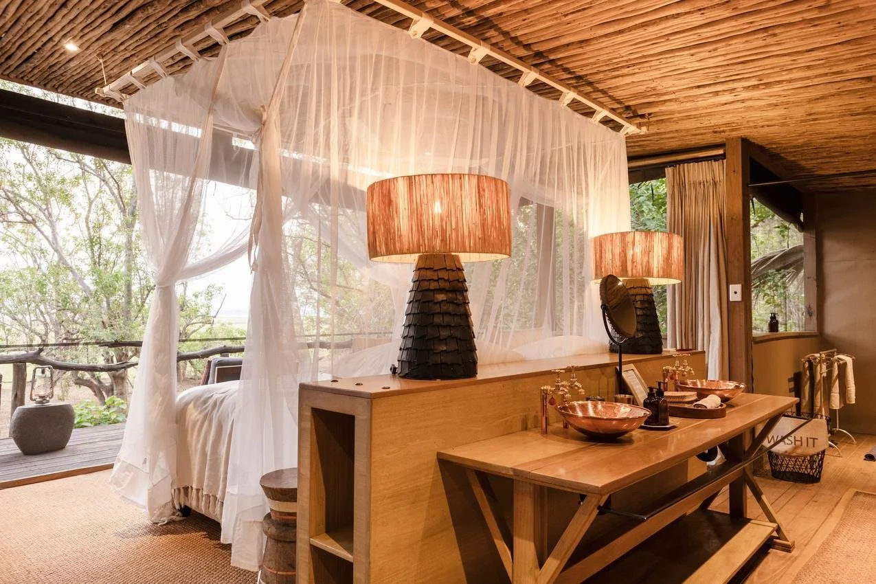 Bedroom with transparent curtains, two large table lamps, a wooden countertop, copper sinks, mirrors, and a sliding glass door leading to a balcony with trees outside.