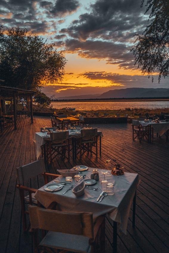 Outdoor restaurant with tables and chairs set for dining, overlooking a river at sunset with dramatic clouds and a mountain range in the distance.