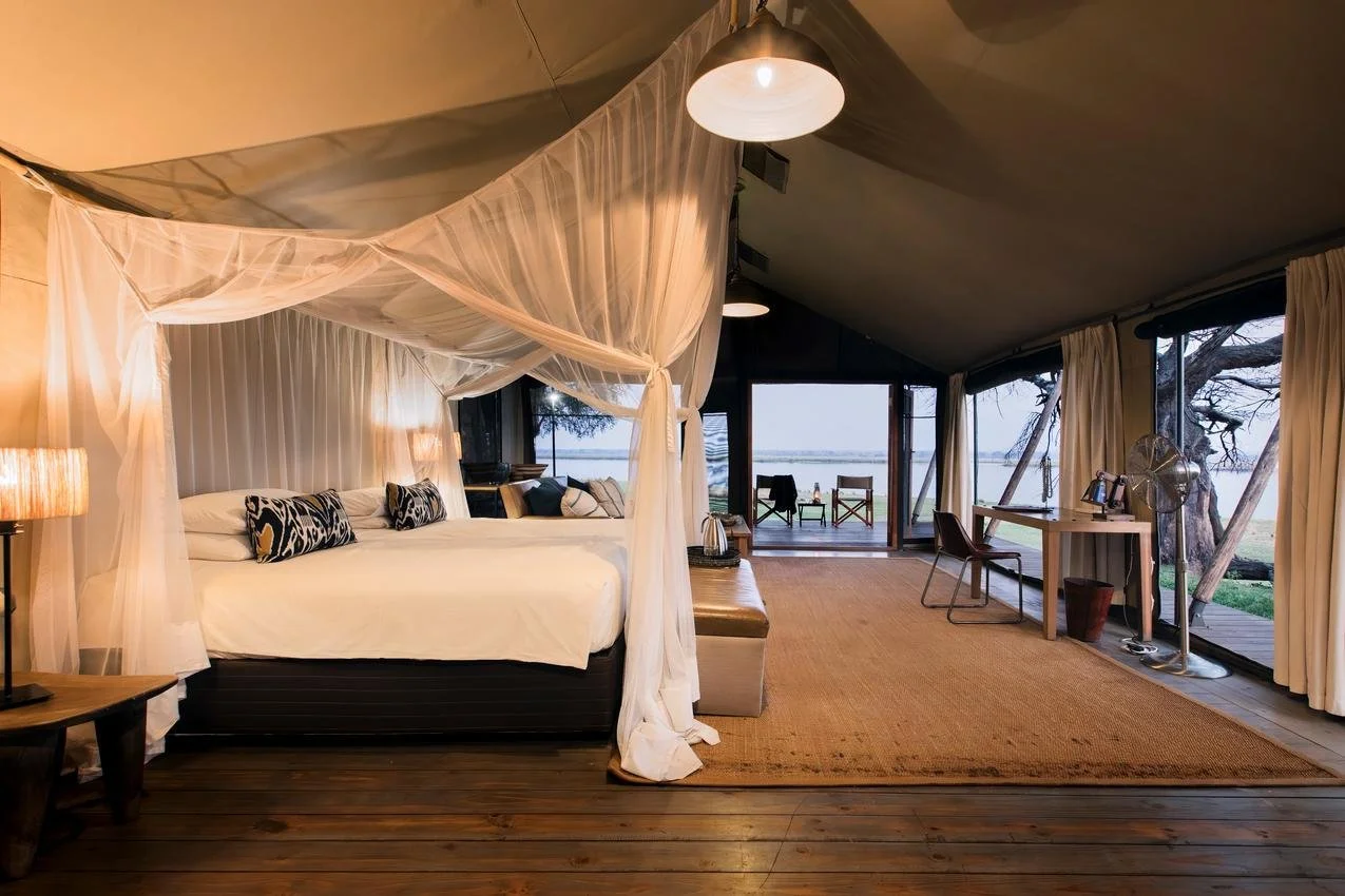 A luxurious bedroom inside a safari tent with a four-poster bed draped in white sheer curtains, a seating area, and large glass doors leading to a view of trees and water outside.