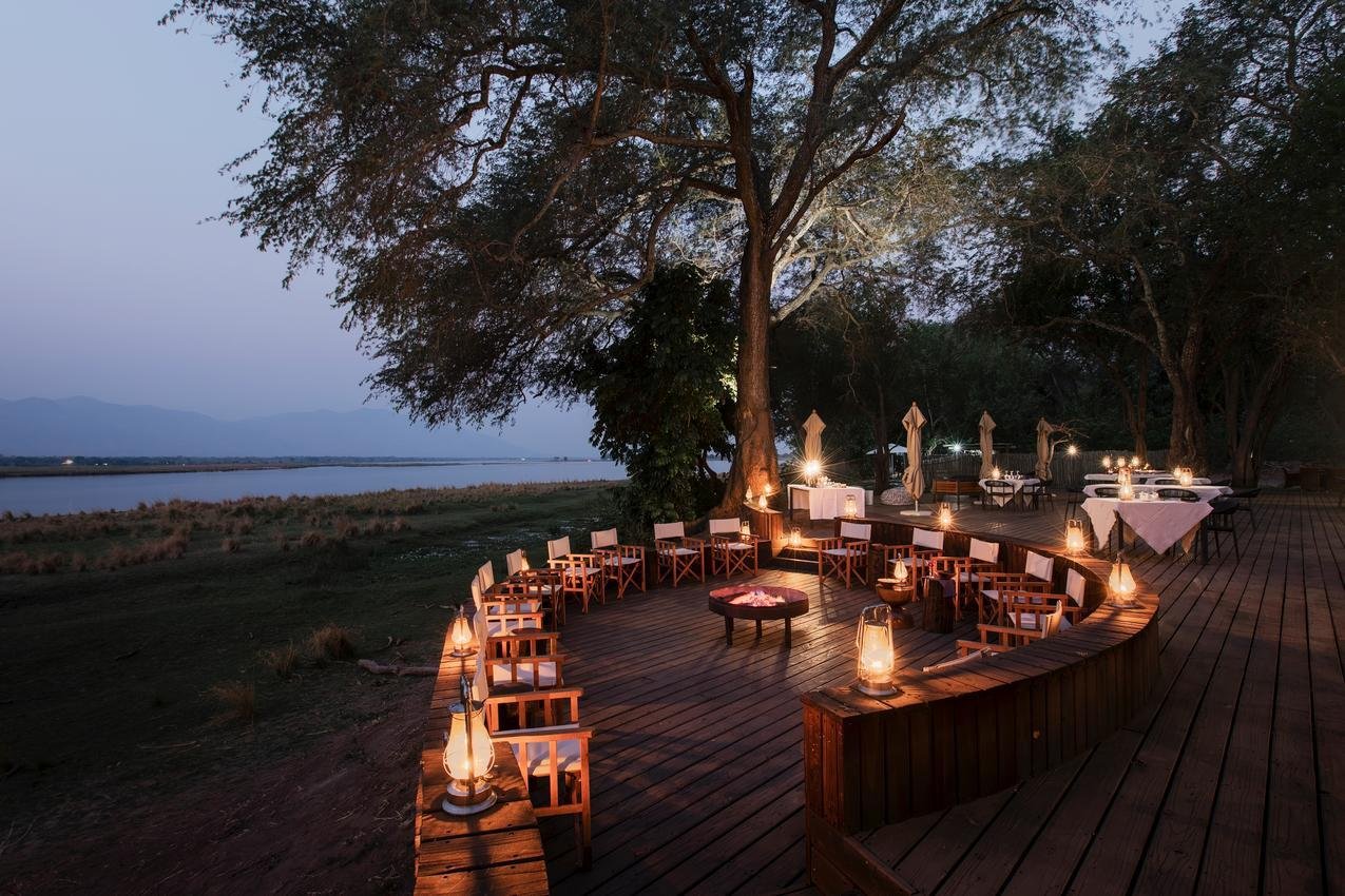 Outdoor dining area on a wooden deck by the water at dusk, with tables, chairs, large trees, and lanterns creating a cozy ambiance.