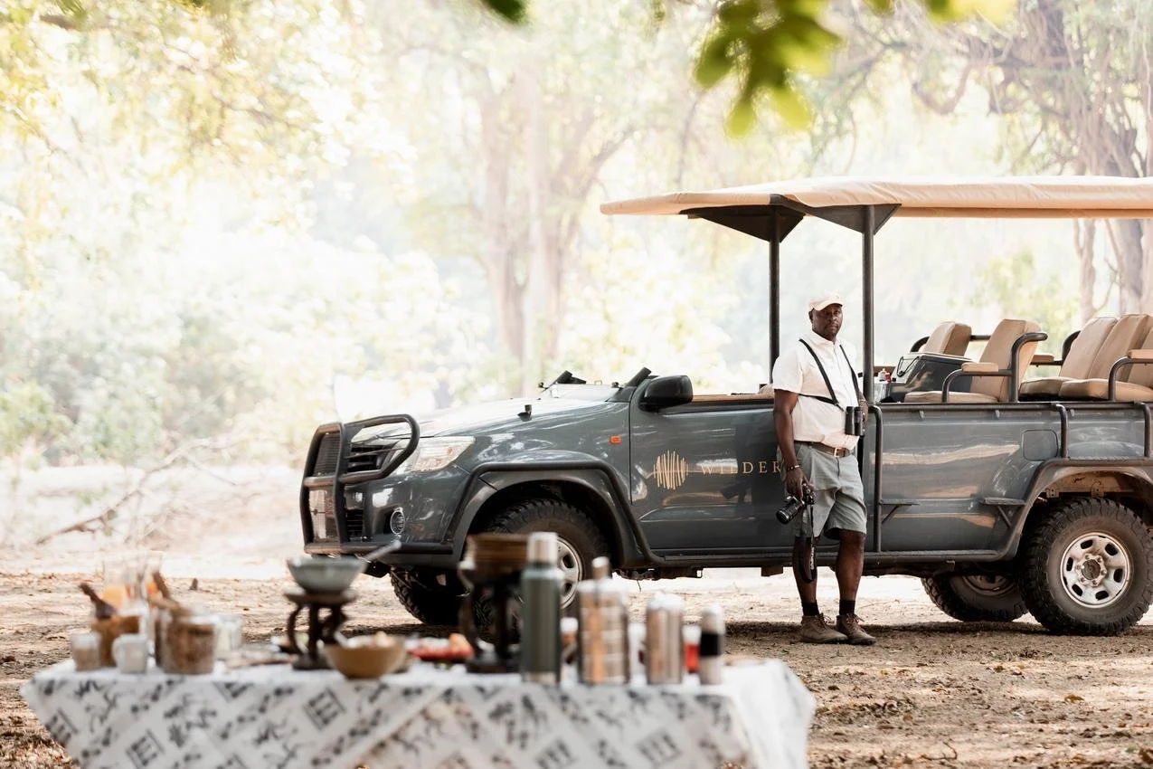 A man standing next to a safari vehicle on a dirt path in a forest, with a table of food and supplies in the foreground.